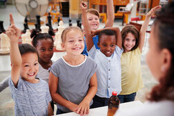 Mädchen und Jungen unterschiedlicher Hautfarben stehen vor einer Lehrperson und melden sich