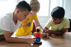 Drei Kinder beugen sich an einem Tisch über einen Spielzeugroboter
