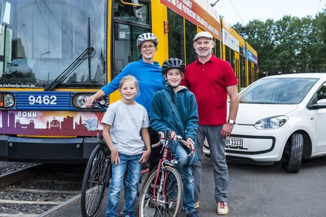 Eine Familie mit Fahrrädern steht vor einem Bus und einem Pkw.