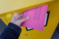 Ein pinker Briefwahlumschlag wird in einen gelben Briefkasten geworfen