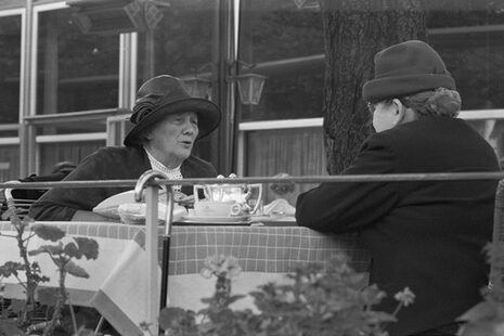 Auf dem Schwarz-Weiß-Foto sind zwei Personen mit Hut und Mütze. Sie sitzen draußen an einem mit Tassen gedeckten Tisch.