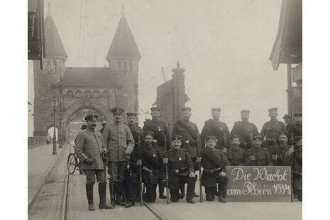 Das Schwarz-Weiß-Foto zeigt Männer in Uniform an einer Brücke. Sie halten ein Schild, auf dem steht: Die Wacht am Rhein. Im Hintergrund ist ein Torbogen der Brücke.