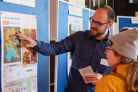 Das Bild zeigt einen Mitarbeitet der Stadtverwaltung, der einer Besucherin an einer Pinnwand ein Plakat zum Klimawandel in der Stadt erläutert.