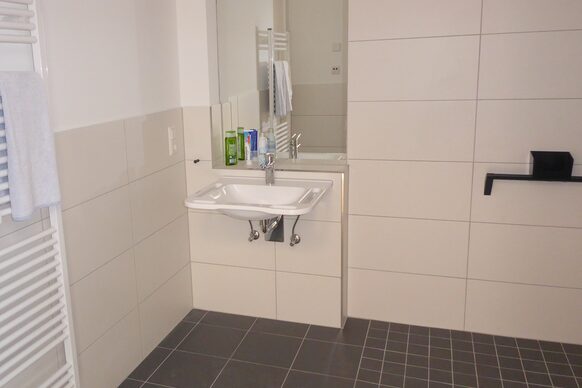 Seniorengerechtes Badezimmer mit cremefarbenen Wandfliesen, Waschbecken, Spiegel und bodengleicher Dusche