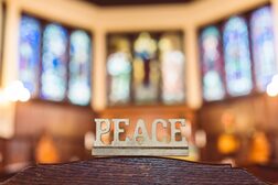 Das Wort Peace als Skulptur in einem Kirchenraum vor bunten Glasfenstern