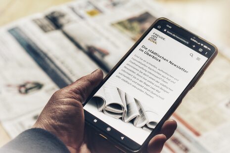 Eine Hand hält ein Smartphone, auf der die Newsletterseite der bonn.de zu sehen ist. Im Hintergrund liegt eine Tagesszeitung.