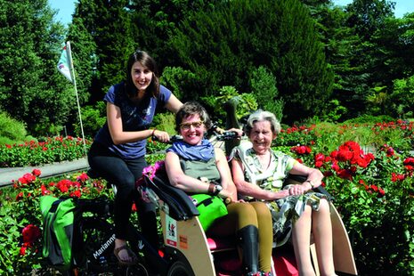 Eine junge Frau fährt mit zwei Seniorinnen mit einer Rikschau durch einen Park.