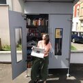 Eine Frau mit einer Zeitung in der Hand steht lachend vor einer geöffneten Freebox
