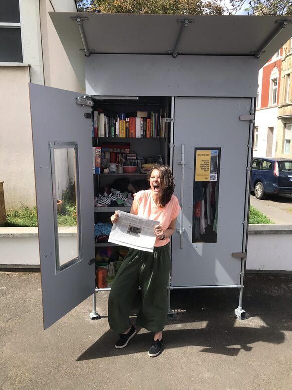 Eine Frau mit einer Zeitung in der Hand steht lachend vor einer geöffneten Freebox