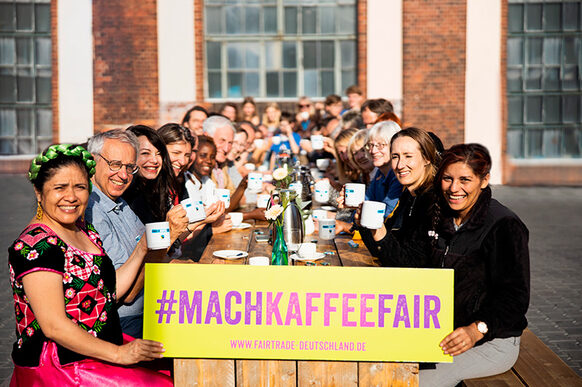 Eine Gruppe Menschen sitzt draußen an einer langen Holztafel und zeigt ein Transparent mit der Aufschrift #Mach Kaffee fair