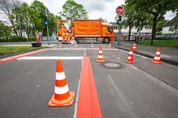Das Bild zeigt die Ringstraße in Beuel, auf der Pylonen stehen und rote Markierungen für die Fahrradstraße aufgebracht sind. Im Hintergrund stehen Mitarbeiter der beauftragten Firma mit ihrem Einsatzfahrzeug.