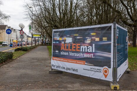 Ein großformatiges Plakat mit der Aufschrift "ALLEEmal einen Versuch wert" macht an der B9 auf den Verkehrsversuch auf der Adenauerallee aufmerksam.