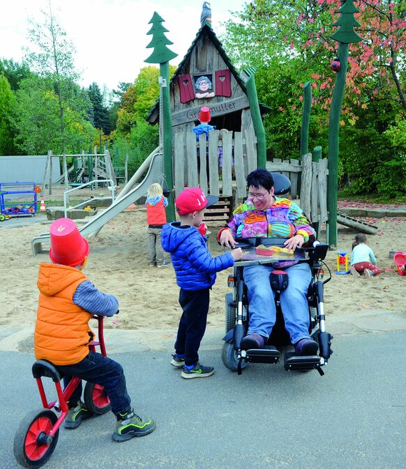 Das Bild zeigt Tanja Rösgen, die in einem Rollstuhl sitzt, und mit Kindern der Kita Am Wolfsbach auf dem Außengelände spielt.
