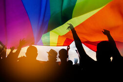 Man sieht die Silhouetten mehrerer Personen, die unter einer Regenbogenfahne tanzen