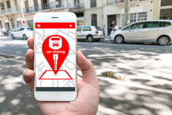 Auf einem Smartphone ist eine Carsharing-App zu sehen, im Hintergrund geparkte Pkw