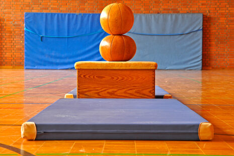 In einer Turnhalle steht ein Holzkasten auf blauen Matten. Oben sind zwei Medizinbälle aufeinandergestapelt.