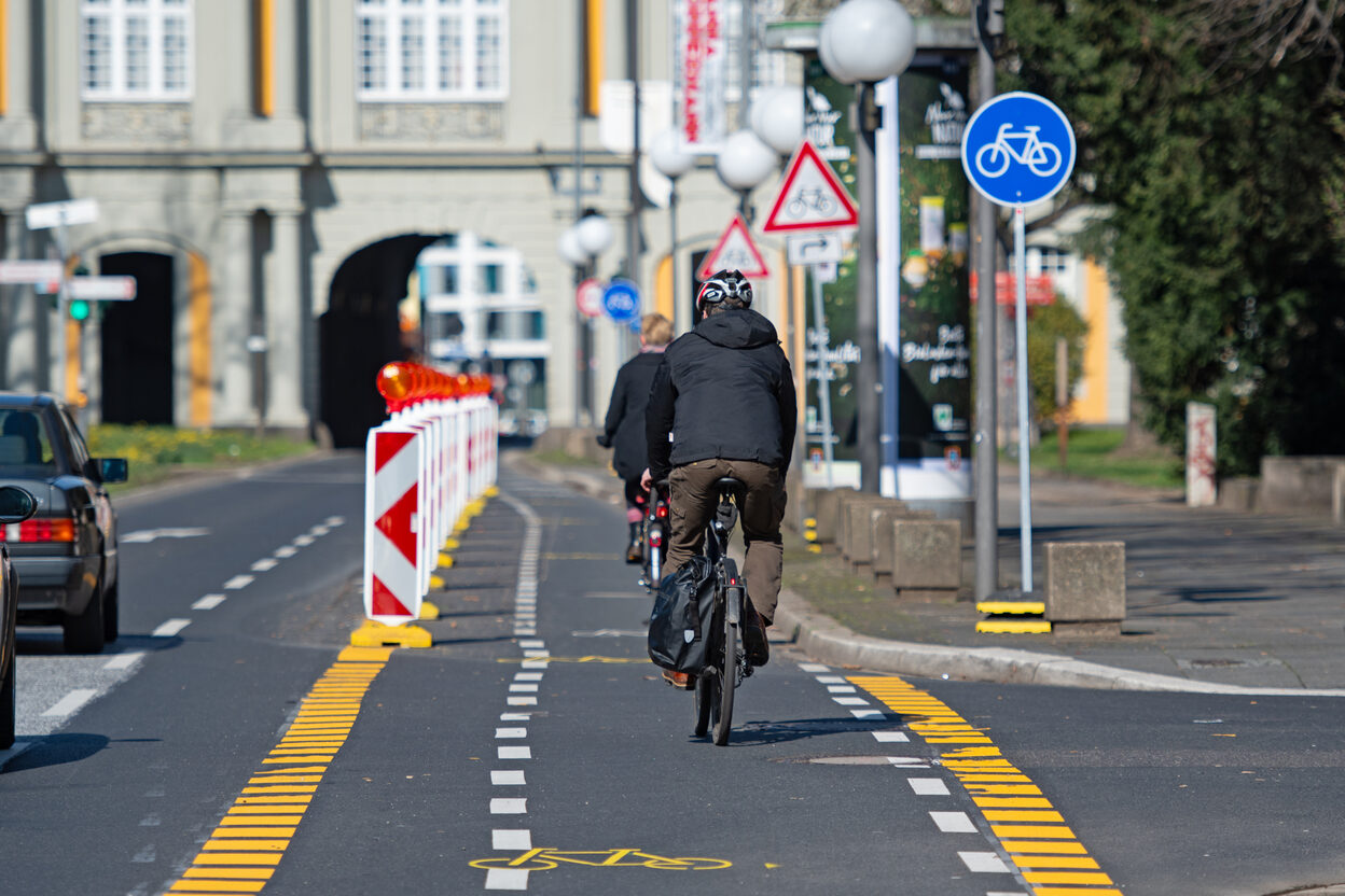 Ein Radfahrer fährt auf der Adenauerallee auf der rechten, für den Radverkehr mit gelben Linien und Baken eingerichteten Fahrbahnhälfte in Richtung Koblenzer Tor. Links wird er von Autos auf der Kfz-Spur überholt.