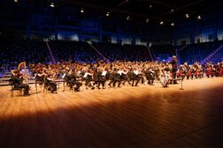 Zu sehen ist ein Orchester in einer beleuchteten Halle mit leeren blauen Sitzplätzen im Hintergrund