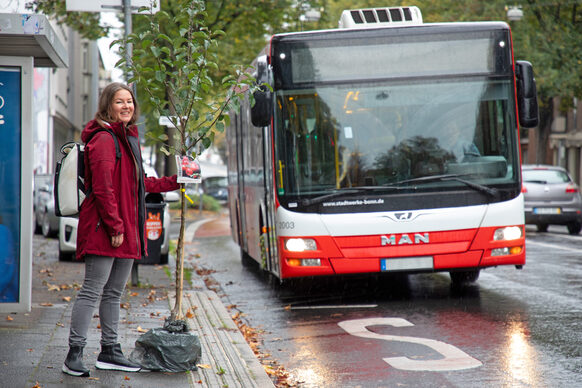 Eine Frau wartet mit einem Obstbaum an der Haltestelle auf den Bus.