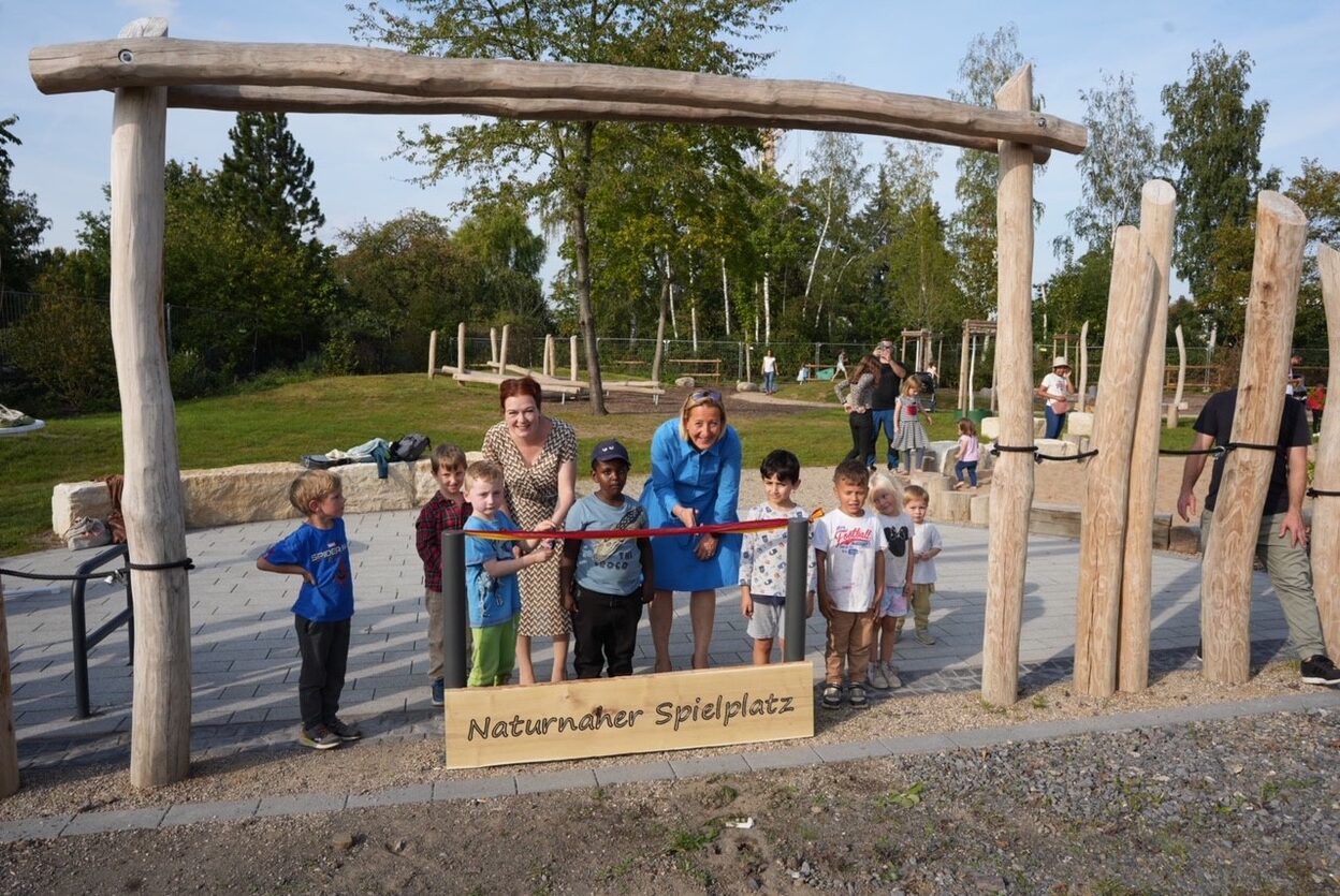 Oberbürgermeisterin und Bezirksbürgermeisterin durchschneiden gemeinsam mit Kindern ein Band auf einem Spielplatz.