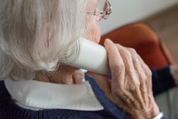 Eine von der Seite fotografierte Seniorin hält einen weißen Telefonhörer in der Hand.