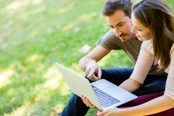 Eine Frau und ein Mann sitzen auf einer Wiese und schauen auf ein Laptop.