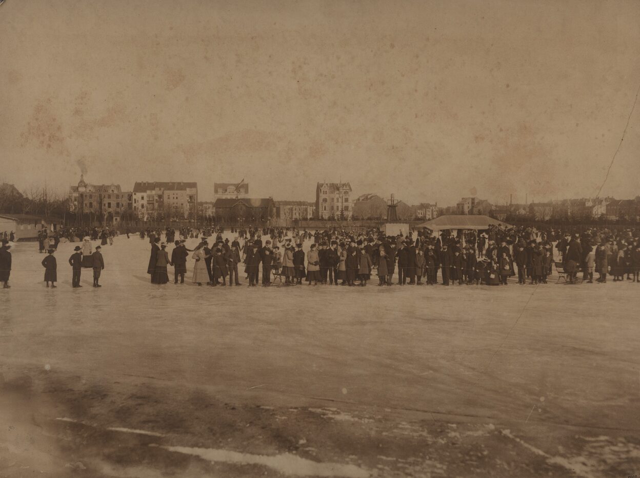 Ein historisches Schwarz-Weiß-Foto zeigt viele Menschen, die auf einem Sportplatz Schlittschuh fahren.