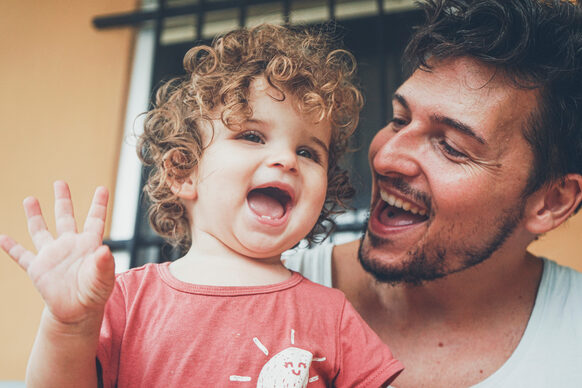 Ein Vater hat ein Kleinkind auf dem Arm und lacht mit ihm gemeinsam