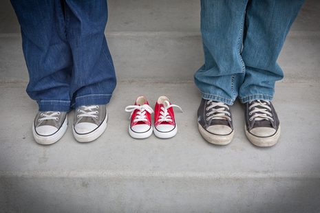 Foto von den Beinen von zwei Personen mit Schuhen. In der Mitte steht ein paar Babyschuhe.