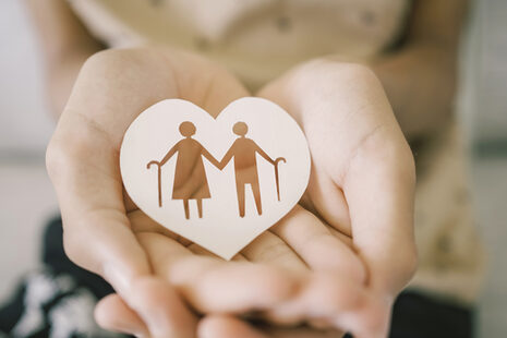 Eine junge Person hält ein Holzschildchen in Herzform in den Händen, auf dem ein Seniorenpaar mit Gehstock zu sehen ist