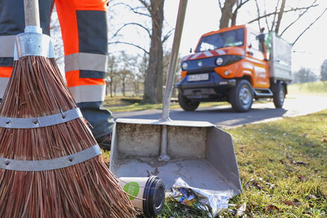 Eine Person in orangener Kleidung der bonnorange AöR in Nahaufnahme kehrt im Vordergrund Abfälle mit einem Besen in ein Kehrblech, im Hintergrund ist verschwommen ein Elektrofahrzeug zu sehen.