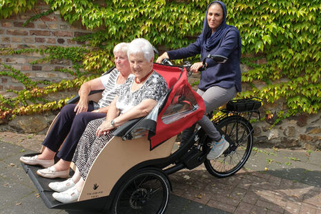 Eine junge Betreuerin fährt zwei Seniorinnen mit einer Fahrrad-Rikscha.
