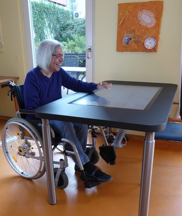 Eine lachende Frau im Rollstuhl sitzt an einem Tisch mit großem Tablet.
