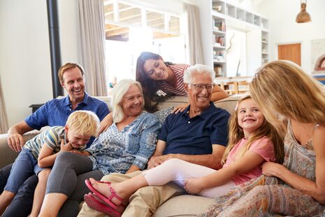 Eine Familie mit drei Generationen Großeltern, Eltern und Jugendlichen sowie Kleinkindern sitzt lachend auf einem Sofa