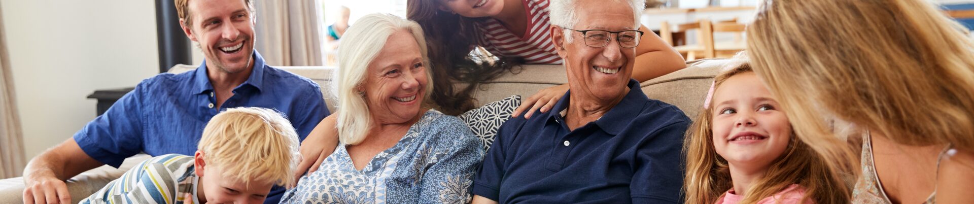 Eine Familie mit drei Generationen Großeltern, Eltern und Jugendlichen sowie Kleinkindern sitzt lachend auf einem Sofa