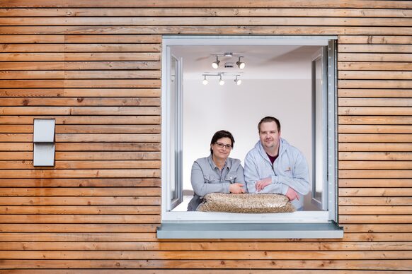 Ein Ehepaar schaut in seinem Holzhaus aus dem Fenster, gestützt auf einen Sack mit Holzpellets