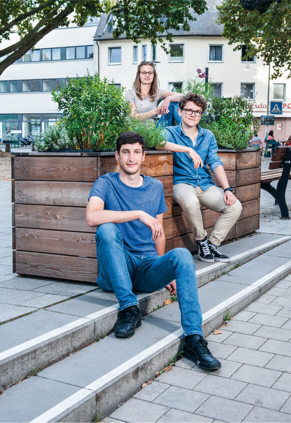 Zwei junge Männer und eine junge Frau sitzen auf einer bepflanzten Holzbank.