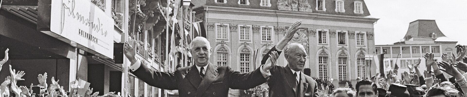 Der französische Präsident Charles de Gaulle und Bundeskanzler Konrad Adenauer fahren 1962 in einem offenen Auto über den Bonner Markt und winken