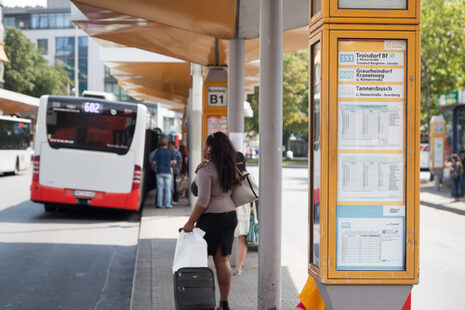 Eine Frau zieht auf einem Bussteig des Zentralen Omnibusbahnhofs einen Koffer hinter sich her; seitlich ist eine Vitrine mit einem Fahrplan zu sehen