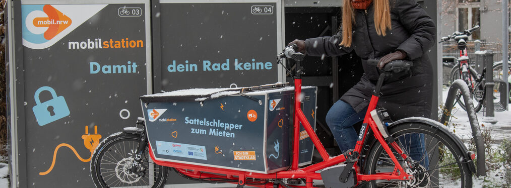 Das Bild zeigt eine Frau mit einem gemieteten Lastenrad vor einer Mobilstation in Bonn.