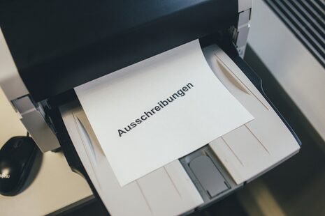 Aus einem Drucker kommt ein Stück Papier mit der Aufschrift Ausschreibungen.