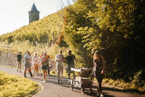Eine Gruppe von Personen läuft mit einem Bollerwagen und Wein die Weinberge entlang.